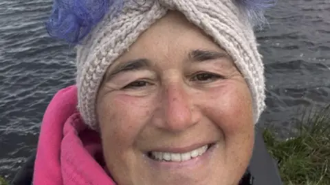 Vicki Attenborough stands in front of a body of water, she wears a pink hoodie under a grey jacket and a cream knitted headband. She has curly purple hair which is pinned on top of her head