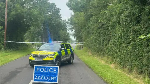 Fermanagh Herald A blue sign in the middle of a road which read POLICE ACCIDENT - trees and grass verge on side of the road. Behind the sign is a police car which blue lights with a tape behind. 