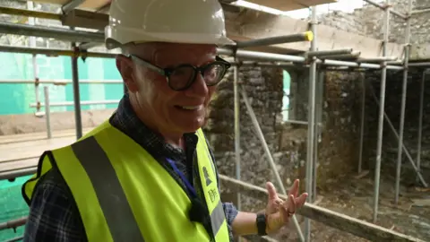 David Conn from the Tavistock Heritage Trust is wearing a hard hat and high-visibility vest. He stands inside Betsy Grimbal's Tower, surrounded by scaffolding and exposed stone walls during restoration work.