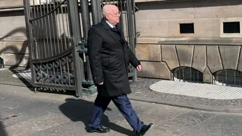 Alex Lawrie A bald man wearing glasses and a dark suit and jacket walks along a street in front of a stone building and metal gate. 