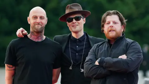 Three men stand side by side smiling at the camera. Mince Fratelli, a bald man with a tattooed neck wearing a black t-shirt, is on the far left. Jon Fratelli is in the centre and wears a brown hat, black sunglasses and a dark suit with earphones dangling on his chest. On the right is Barry Fratelli who has brown hair and a ginger beard and wears a dark jacket with his arms crossed across his chest.
