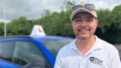 A man with a short grey beard stood in front of a blue car with a learner driver sign on the top of it. He is wearing a beige cap with sunglasses on his head and a white polo top which has the words 'able automatic driving school' stitched on it. He is smiling and looking right at the camera. 