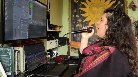 Lily sings into a wired microphone at a desk in her bedroom. In front of her are a laptop and screen with editing software. Lily is wearing a black shirt and purple and red scarf.