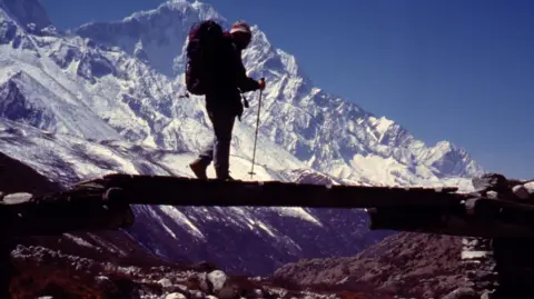 Caradoc Jones A man in mountaineering kit, with a backpack, walking along a ledge holding a walking pole with snowy mountains behind him.
