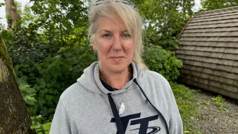 Sam Jones wears a grey hooded jumper, you can see the top of two embroidered letters which read TT in black. She wears a necklace in the shape of the Isle of Man. She has blonde hair which is tied up, and sunglasses on her head. She stands next to some green bushes.