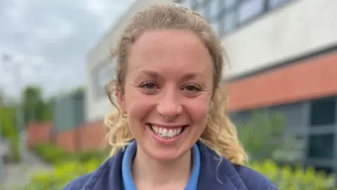 A woman with blonde curly hair that's tied back in a ponytail is smiling happily into the camera. She is wearing a blue district nurse polo shirt with a blue fleece over the top. The NHS logo is visible on the left hand breast pocket.