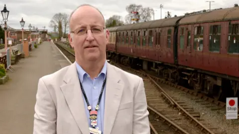 A man in a white suit and blue shirt and also wearing glasses and a lanyard is looking at the camera. He is on a platform at a railway station.