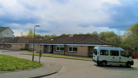 NHS Lothian Exterior of a brown bungalow with a white van outside. It is the end of a street with a road that curves around it. There are blue, cloudy skies.