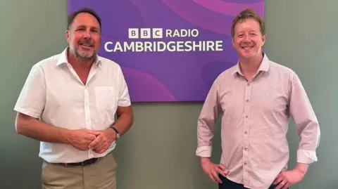 Charlotte Allen/BBC Two men smile and stand either side of a purple sign that says 'BBC Radio Cambridgeshire' 