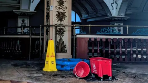 The inside of the Kursaal, a dimly lit atrium with deteriorated flooring. In the foreground is a yellow "caution" cone with two red buckets and a inflatable paddling pool being used to catch dripping water. 