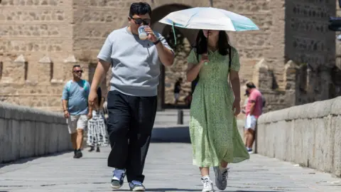 Two people walking, one is drinking some water and one is holding an umbrella