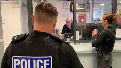 Thames Valley Police Two police officers, one male and one female, book a suspect into custody. They have their backs turned to the camera - with the suspect obscured. A custody sergeant is sitting at a desk behind glass. He is wearing glasses.