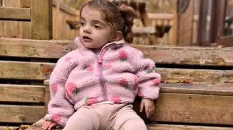 A little girl in a pink coat sits on a wooden bench