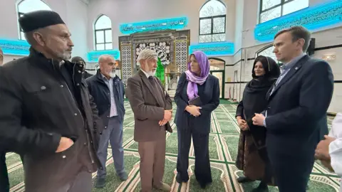 Emma Baugh/BBC Members of the community, alongside the minister, a woman with a purple headscarf and black outfit, standing inside the Mosque with green carpet. Also seen in the pic is Abdul Chaudhuri next to her, Shabina Qayyu, a woman and Andrew Pakes, the city MP.