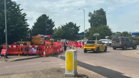 Jaimielee Rendall/BBC Red barriers and road closed signs at a roundabout on which there is a queue of cars.