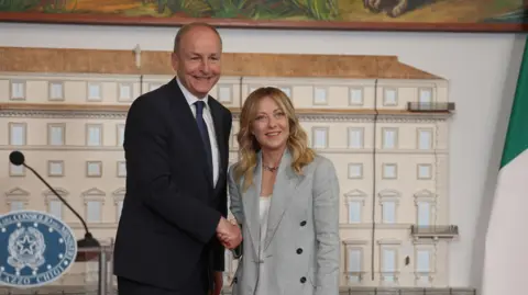 PA Media A man with short brown hair shakes the hand of a woman with long dark-blonde hair. The man, standing on the left, wears a black suit, white shirt and navy tie. The woman wears a grey suit with a white top. 