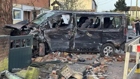 Luke Deal/BBC A dark coloured van badly damaged with dents and scrapes. It has crashed through a brick wall and there are bricks lying across the road.