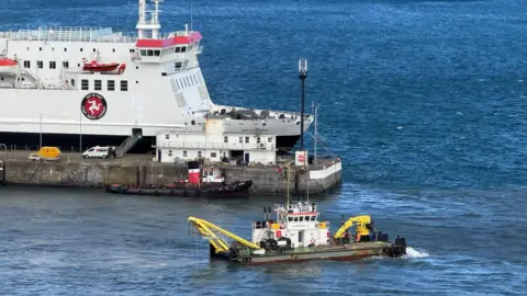 A dredging boat, it has two long yellow cranes going into the water, you can see a ferry moored in the background on a clear day.