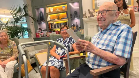 man in a checked shirt smiles broadly as he holds a card, seated in a care home lounge. Behind him a woman in a blue dress smiles, with other guests and a member of staff nearby