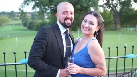 Joe Mintz and Vanessa Curto standing in front of black railings with a golf putting green and trees behind them. Joe is wearing a black suit, a white shirt and black tie. He has a shaved head and a dark beard. Vanessa is wearing a blue dress and a piece of snake jewellery on her upper arm, and has long brown hair tied back. Both are holding champagne flutes and are smiling at the camera.