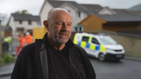 Simon Woodward, who is bald with a beard, stands outside with a police car in the background