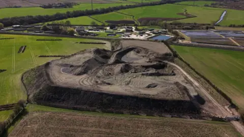 A large waste storage site sits surrounded by fields, next to sewage treatment plant. It looks like a large, raised mound of soil.