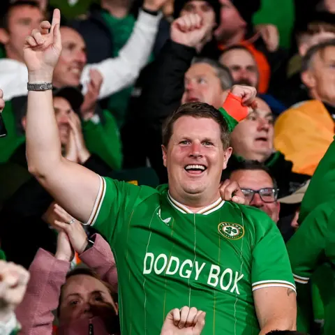 Getty Images A football fan in a green Ireland top is smiling at the camera and pointing his finger in the air. He is standing in front of a busy crowd of supporters. His jersey says 'DODGY BOX' on it where the usual sponsor Sky would be.