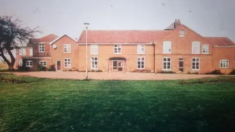 Shirley Oaks Survivors Association Front elevation view of Red House at Buxton, Norfolk, featuring a range of red brick buildings and ancillary buildings with white windows, tiled roofs and gabled ends. There is a lawn and hardstanding in front of it, and a lamp post