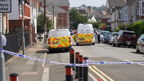 A police cordon can be seen in the forefront. Behind it is a residential road with two police vans parked up.