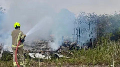 A firefighter pictured from behind as they spray water on to the fire. Smoke billows up from piles of burnt wood on the floor. The fire is in a rural setting with bushes and trees.