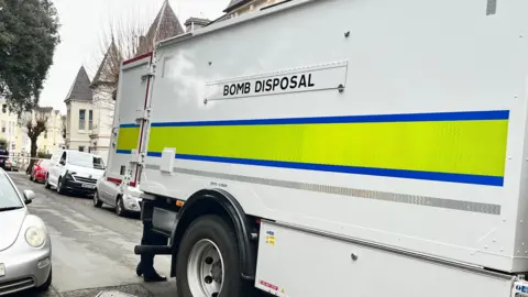 A bomb disposal van. It is a white van with a high visibility green and blue strip on the side. There is police tape in the background. There are cars parked on the road.