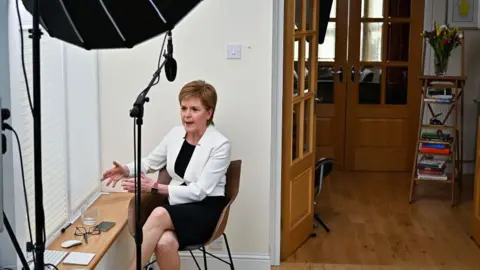 PA Media Nicola Sturgeon sitting on a chair next to the window in her house. She is wearing a white jacket and black dress. The library ladder is in the background. 