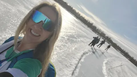 Annette Mawby, a woman wearing green glasses and standing on snow while taking a selfie - with more people seen in the background of her picture.