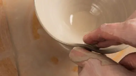 BBC A pottery worker is working on a piece of clay. Fingers can be seen shaping the clay, which is light brown in colour.