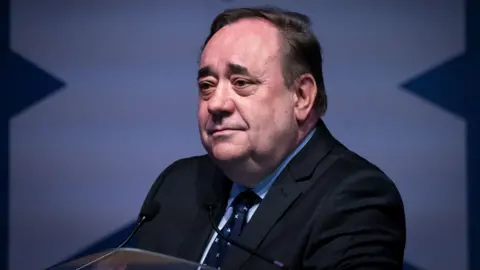 Alex Salmond is sitting looking to the left of camera. He is wearing a dark blue suit, light blue shirt and dark blue tie. He is smiling. There is a saltire in the background.