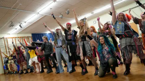 A group of people in a variety of costumes rehearse in a room with a wooden floor. 