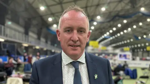 Cathal Mallaghan. He has short light brown hair, wearing a navy blazer, white shirt with blue polka dots, navy tie and Sinn Fein pin on his lapel. 