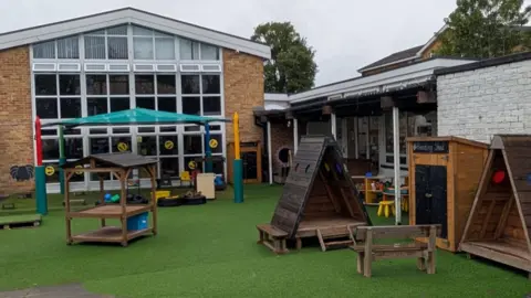 St Peter's School The back of St Peter's School and its playground, which has been partly covered in green astro turf. There are various wooden boxes and triangles around as play areas. The school building has a lot of windows.