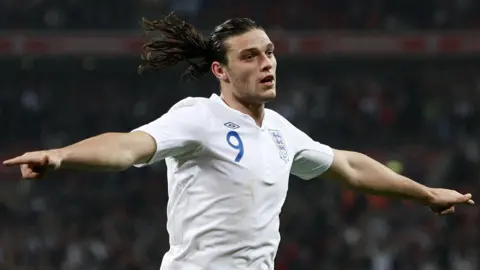 PA Media Andy Carroll is celebrating after scoring a goal for England. He has is arms out by his sides and is wearing the white home shirt with a blue number nine on his chest. Carroll has long brown hair in a ponytail.