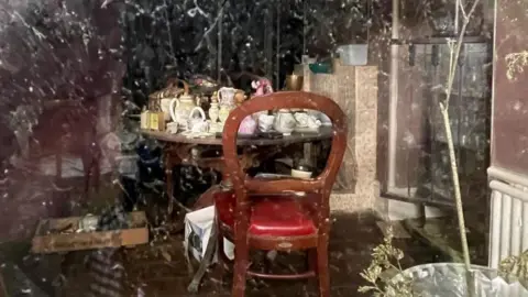 Nick Le Huray An old fashioned table covered in crockery. The photo is taken through a window