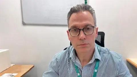 Surgeon Matt Smith has short grey hair and wears dark-rimmed glasses and a blue-and-white striped shirt. He sits in an office with white walls and a whiteboard.