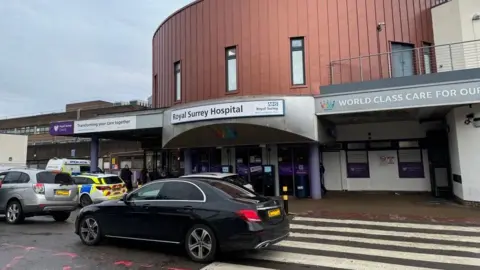 The outside of the Royal Surrey Hospital in Guildford. A sign says 'Royal Surrey Hospital.' Another sign shows the beginning of the slogan 'WORLD CLASS CARE FOR OUR COMMUNITY.' Cars drive across a zebra crossing in front of the hospital.