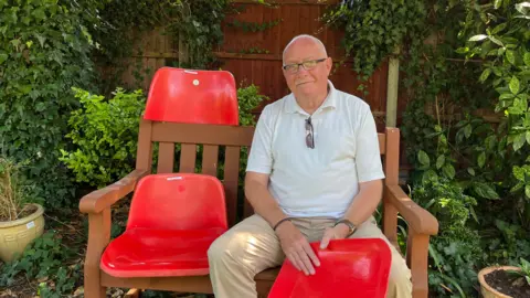 BBC / Patrycja Boryka A bald man with glasses and a grey moustache sitting on a wooden garden bench holding a part of a plastic red seat in his hands, the other parts of the seats are placed to the left of him on the bench and one is on the back of the bench. He's wearing a short sleeve white polo top with a pair of sunglasses hanging on the collar. He's wearing beige chinos and there's a lot of green garden plants and a wooden fence behind him.