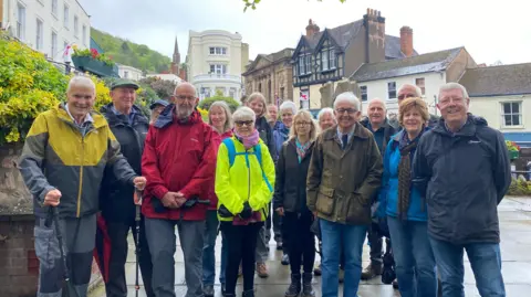 Malvern Civic Society A group of about 16 walkers in a town centre. They are wearing coats or jackets and are all facing the camera with most people smiling. Behind them the tops of buildings, including a church steeple, are visible. 