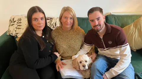 Two women sat beside a man on a green sofa with a dog and various bits of paperwork.