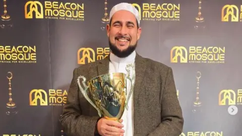 Family photograph Imam Adam Kelwick who has a dark brown beard has his eyes shut as he has a huge smile holding a trophy at the Beacon Mosque Awards 