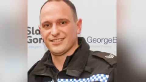 Sgt Paul Williams in his uniform standing in front a sign showing logs for sponsors of the bravery awards.