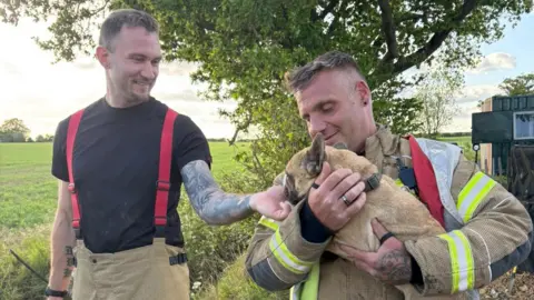 Adriana Hudson A firefighter holds Moose the dog in his arms after he was rescued from a pipe. Another firefighters stands to the side and strokes Moose.