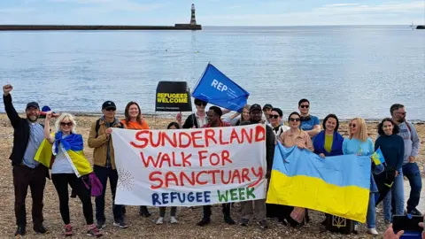 About 20 people holding Refugees Welcome sign, an Ukrainian flag and a Sunderland Walk for Sanctuary Refugee Week flag at Roker Beach in Sunderland. Roker Pier and lighthouse are visible in the background and there is a single boat in the calm blue sea. 