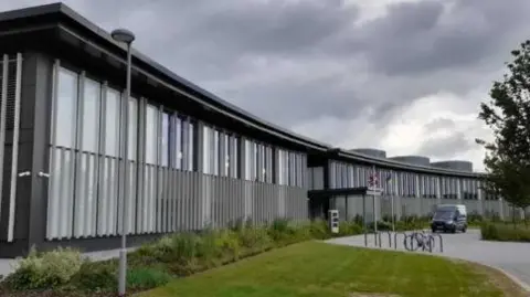 LDRS A modern curved grey and black building, with grass, shrubs and a road to the front.